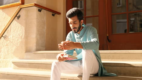 Handsome-young-Hindu-man-in-traditional-clothes-sitting-on-steps-outdoors-and-reading-notes-in-notebook.-Male-from-India-checking-planner.-Day-planning.-Diary.